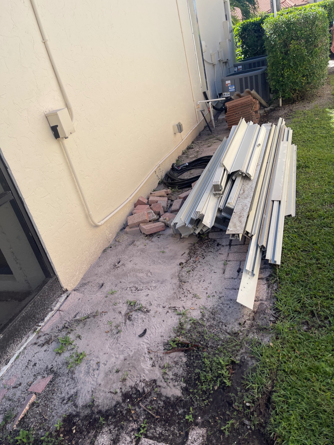 Removed aluminum hurricane shutter panels, brick pavers and roof tile stacked along the side wall of a South Florida home, ready for All Out Rubbish to haul away