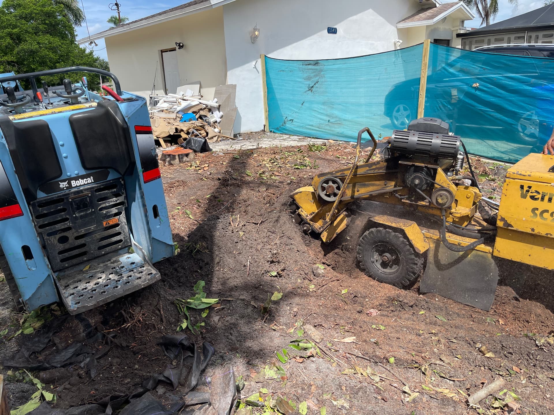 Bobcat compact loader beside a stump grinder working on a cleared lot with construction debris staged for removal