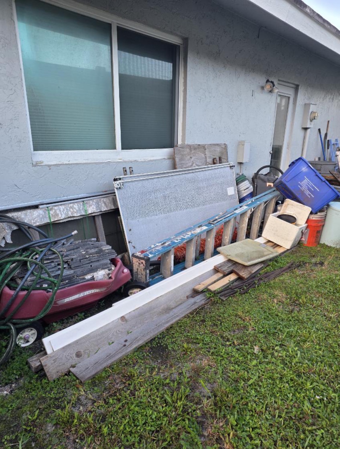 Before and after junk removal: construction tiles, lumber, buckets and a ladder cleared from against a house wall by All Out Rubbish (before)